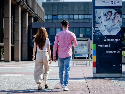 Zwei Personen gehen über den Campus der Ruhr-Universität Bochum an einer Willkommenstafel vorbei.
