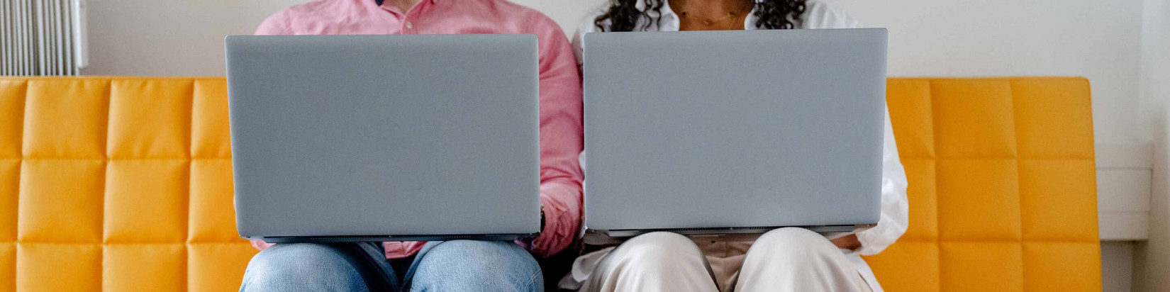 Two people sit next to each other on a yellow sofa and work on their laptops.