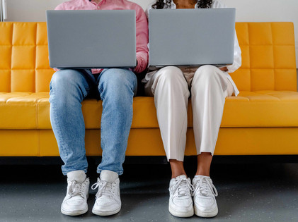 Zwei Personen sitzen nebeneinander auf einem gelben Sofa und arbeiten an ihren Laptops.